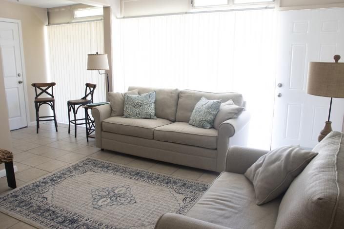 Bright living room with beige sofas, patterned rug, chairs, floor lamps, and tall windows with white blinds