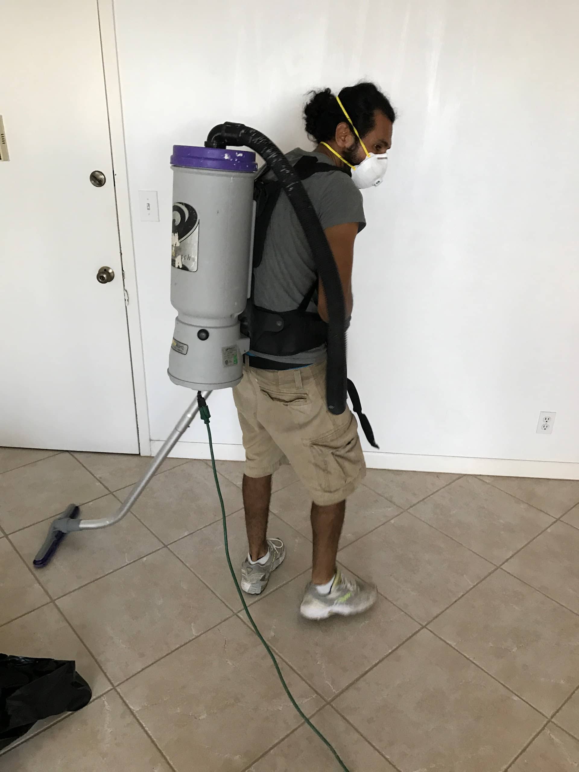Man wearing a backpack vacuum, respirator, khaki shorts, and sneakers. Indoors, white walls, tile floor.