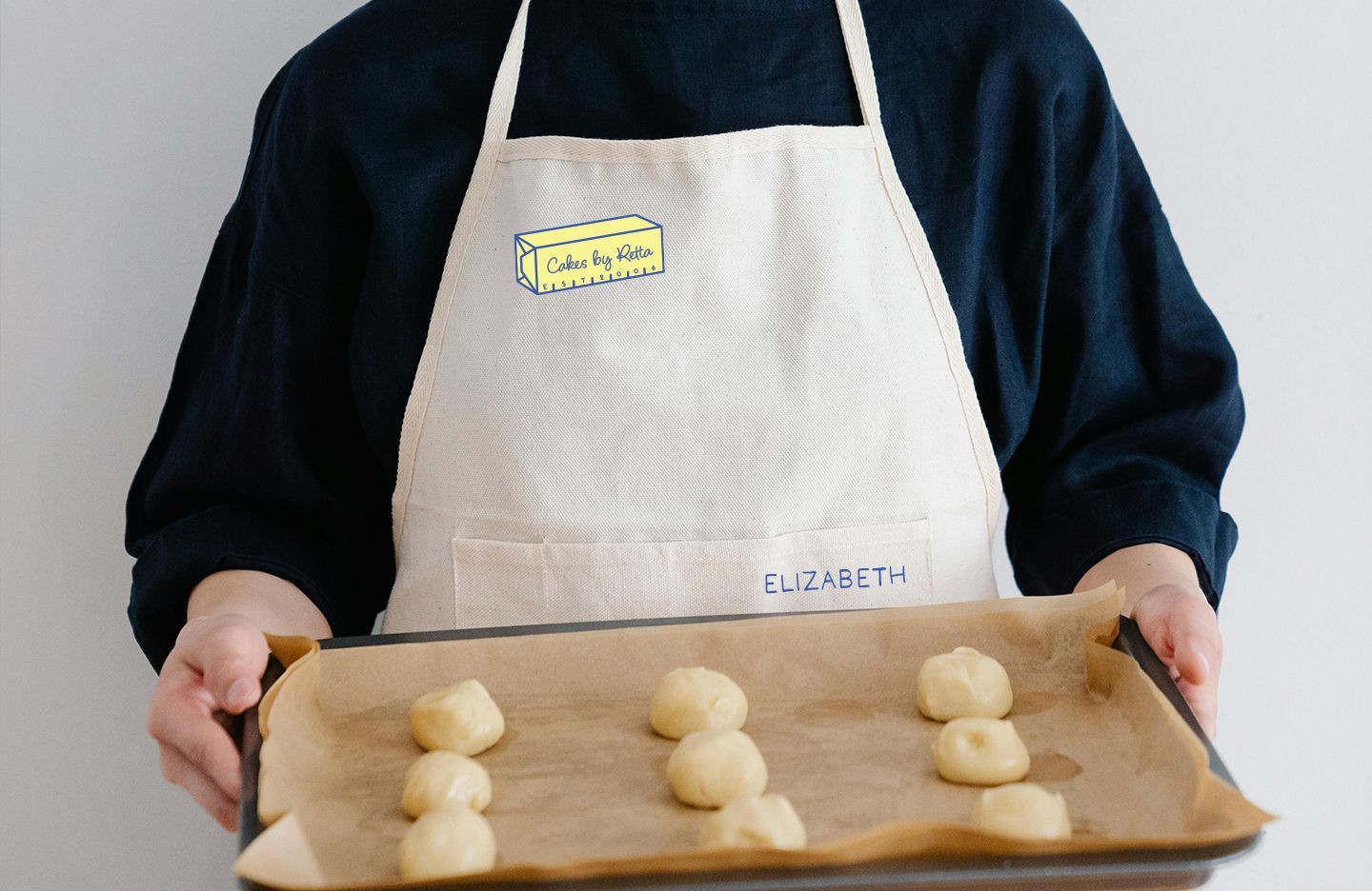 Person holding a baking sheet with raw cookie dough balls, wearing an apron with 