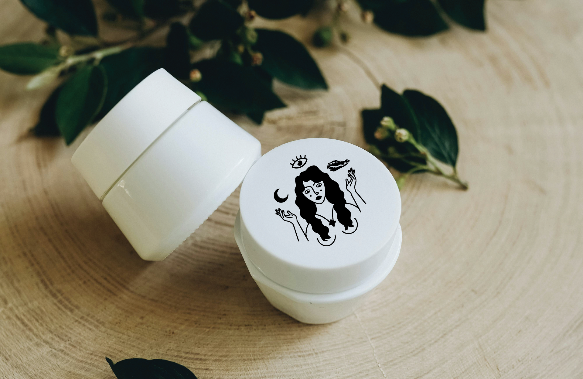 Two House of Wayward Women white cosmetic jars, one open, on a wooden surface with greenery.