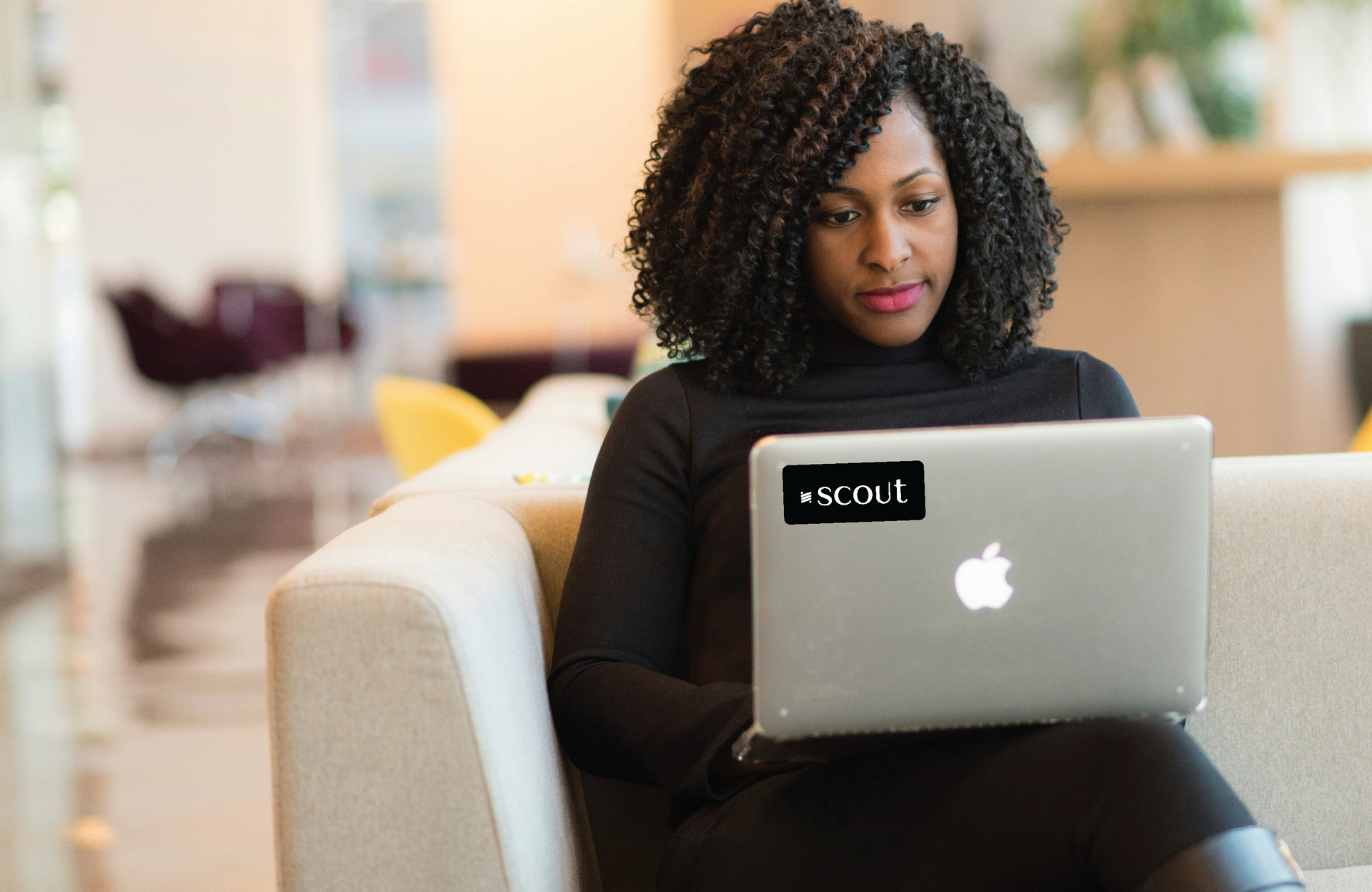 Woman with curly hair works on a laptop, in a lobby setting. Black turtleneck and sticker