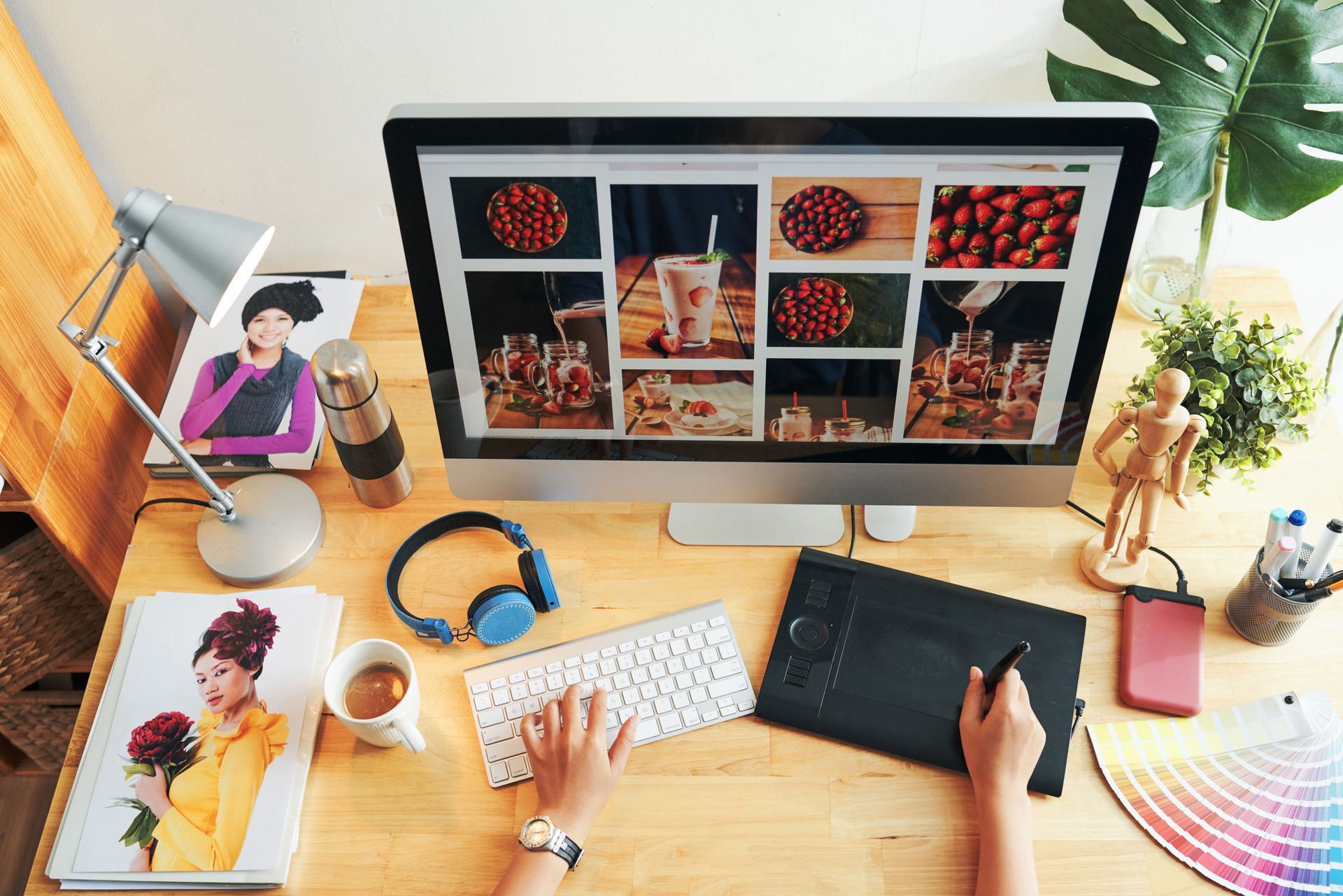 A person uses a computer, tablet, and keyboard at a desk. The computer shows a photo grid.