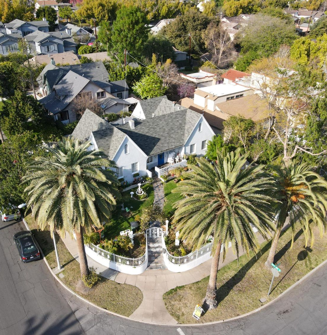 An aerial view of a light blue house surrounded by palm trees and greenery on a corner lot.
