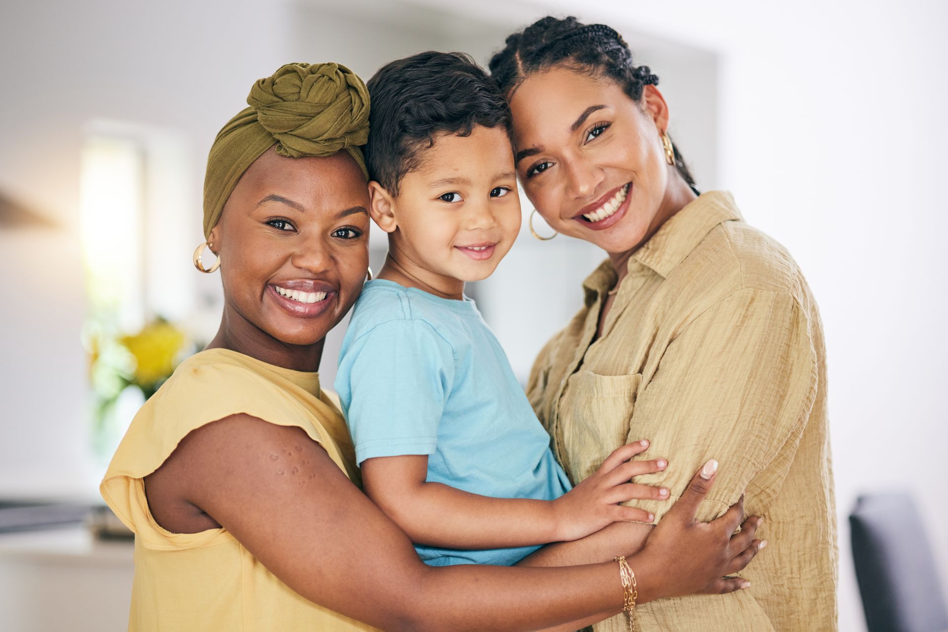 Three smiling people hugging indoors, wearing light-colored clothes.