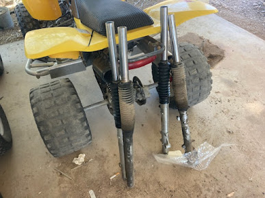Rear view of a yellow ATV with exposed front suspension and large knobby tires on a dirt floor.
