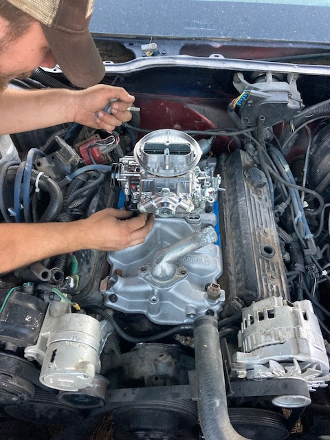 Mechanic working on a car engine in an open hood bay, with tools and exposed engine components.