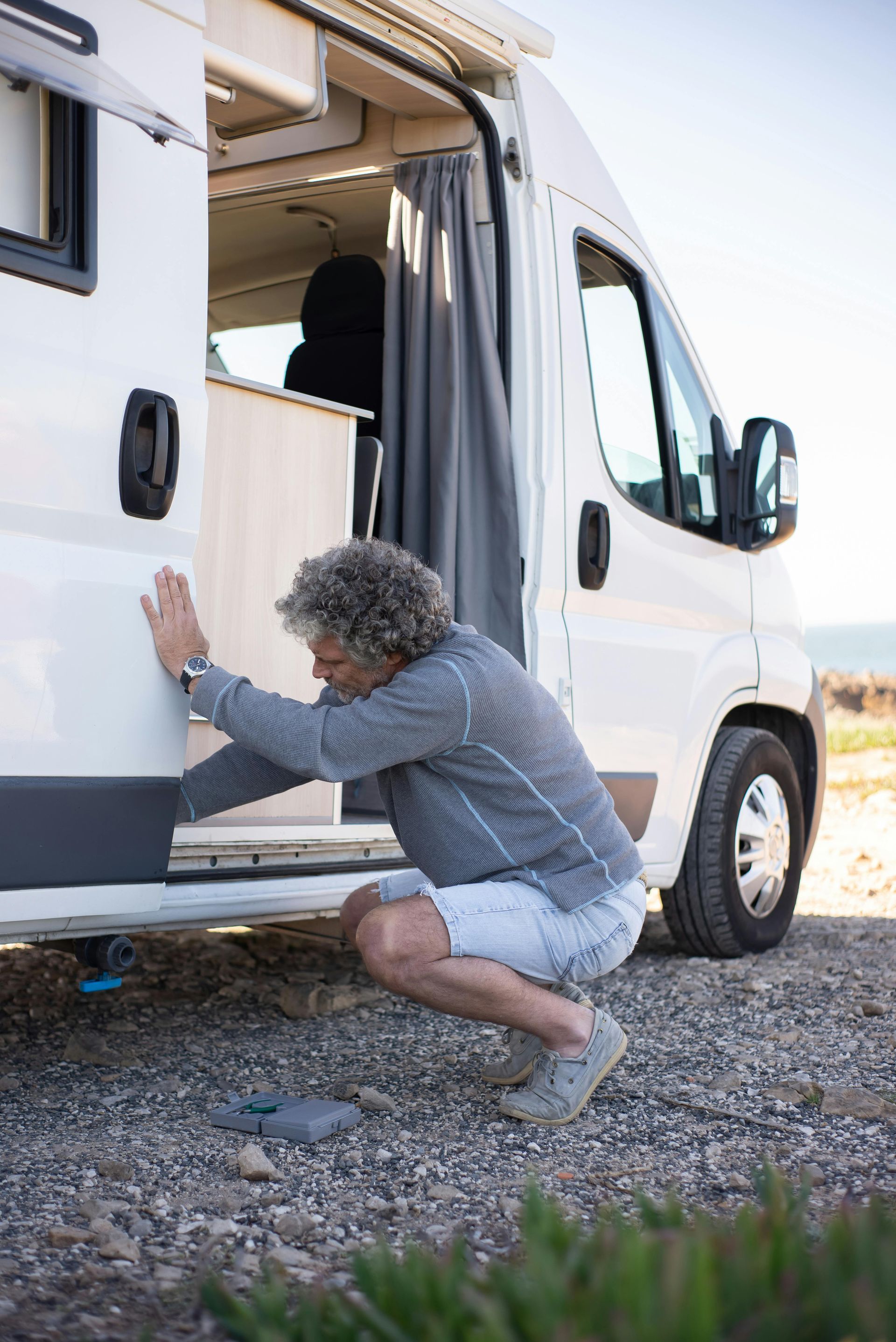 Person crouching beside a white camper van, inspecting or opening the side door on a gravel roadside.