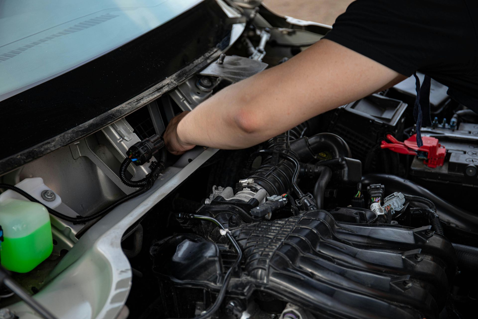 Person working under a car hood in an engine bay with exposed components and a green fluid reservoir