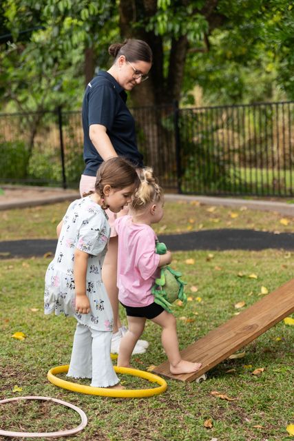 Adult supervising two children using a toy hose on a lawn, with hoops and a ramp in a park