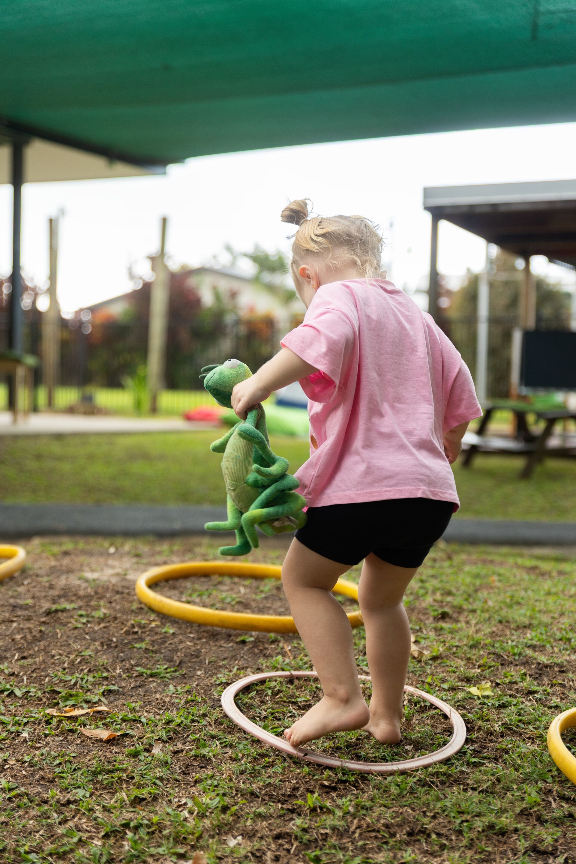 Child in a pink shirt and black shorts plays with a green toy snake on stepping rings in a park.