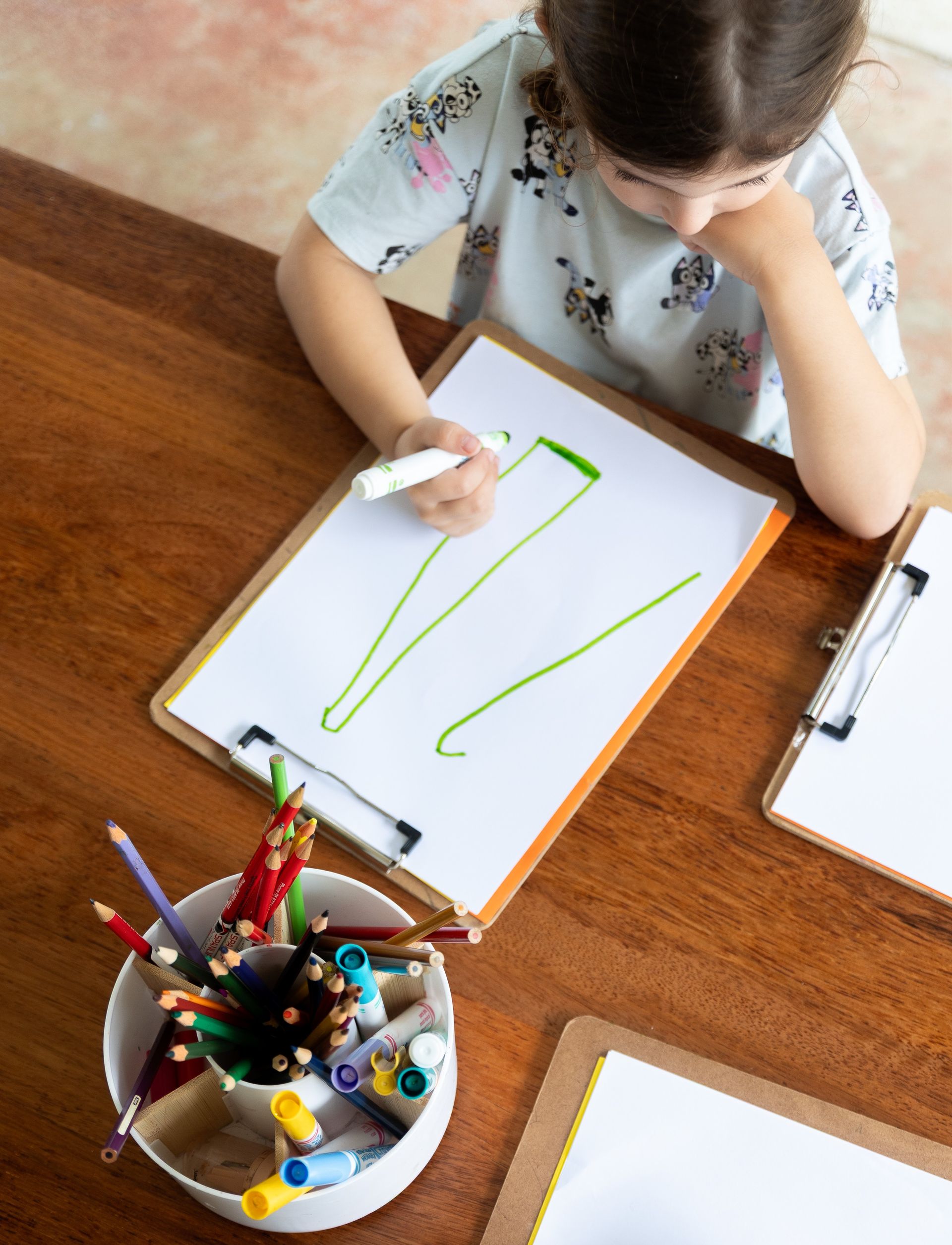 Child drawing green lines on a whiteboard at a wooden table, with art supplies nearby