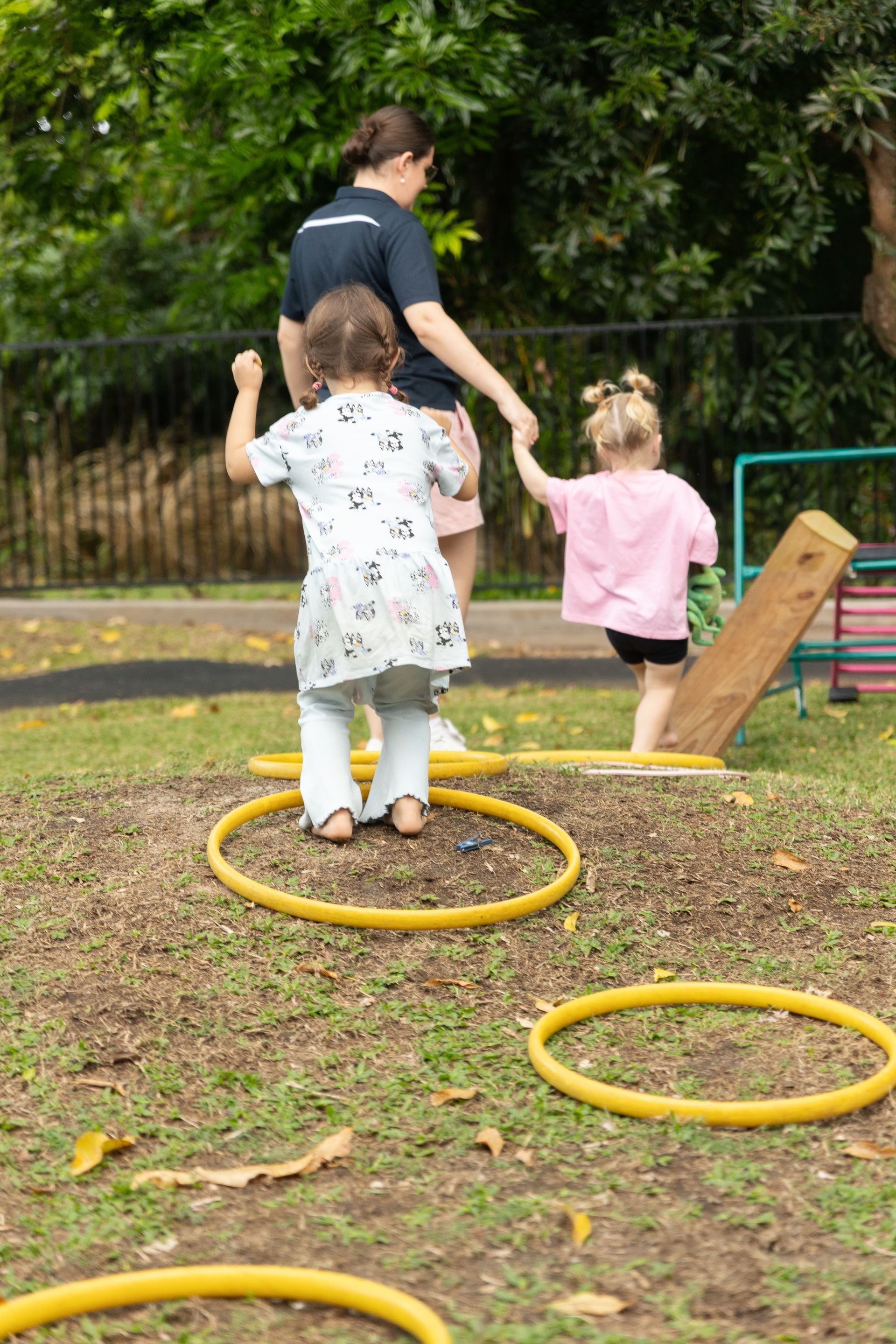 Two children and an adult playing on yellow hoops in a park, with trees and a bench nearby.