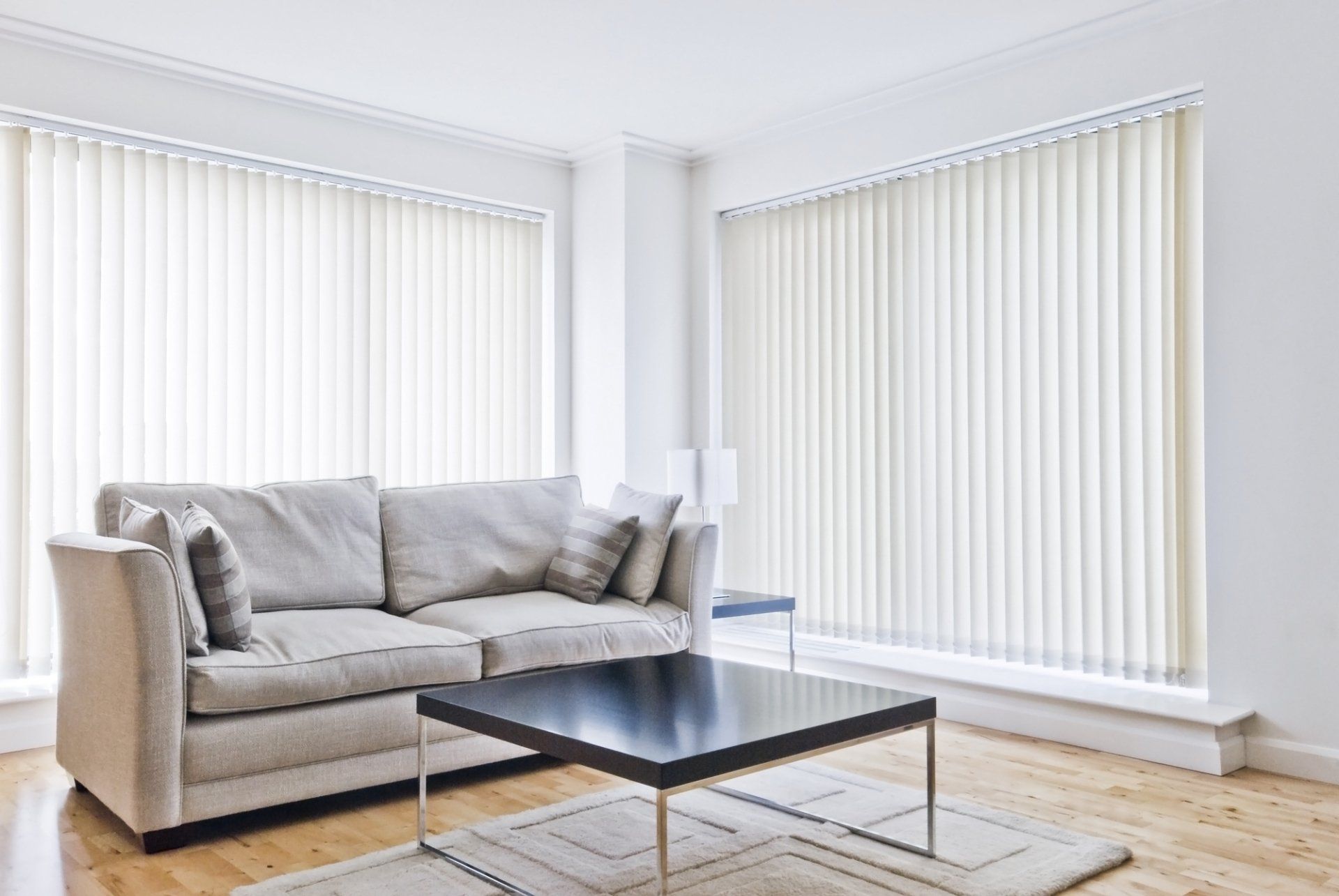 Closed vertical blinds on each wall of loungeroom. Grey couhc on timber floor, behind black coffee table on rug.
