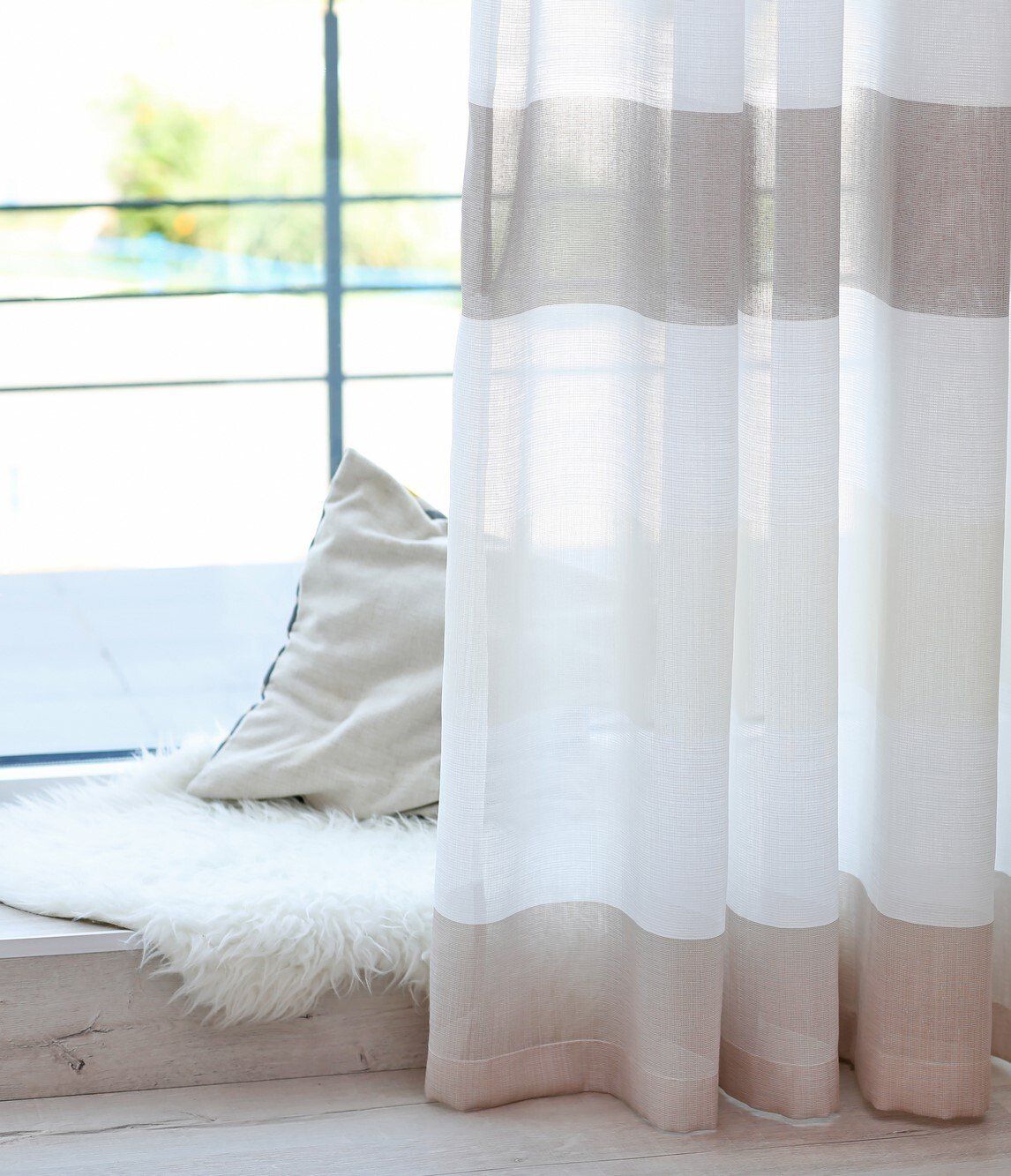 Close up of mocha and white curtain pulled open to show sunny outdoor view through windows with rug and cushion.