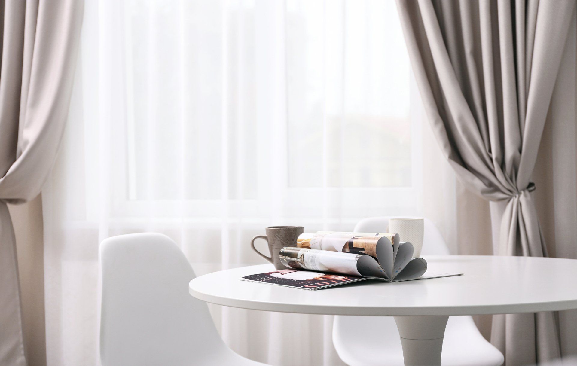 Kitchen with soft curtains open showing white sheers. White round table and chairs in foreground, with open magazine and coffee mugs.