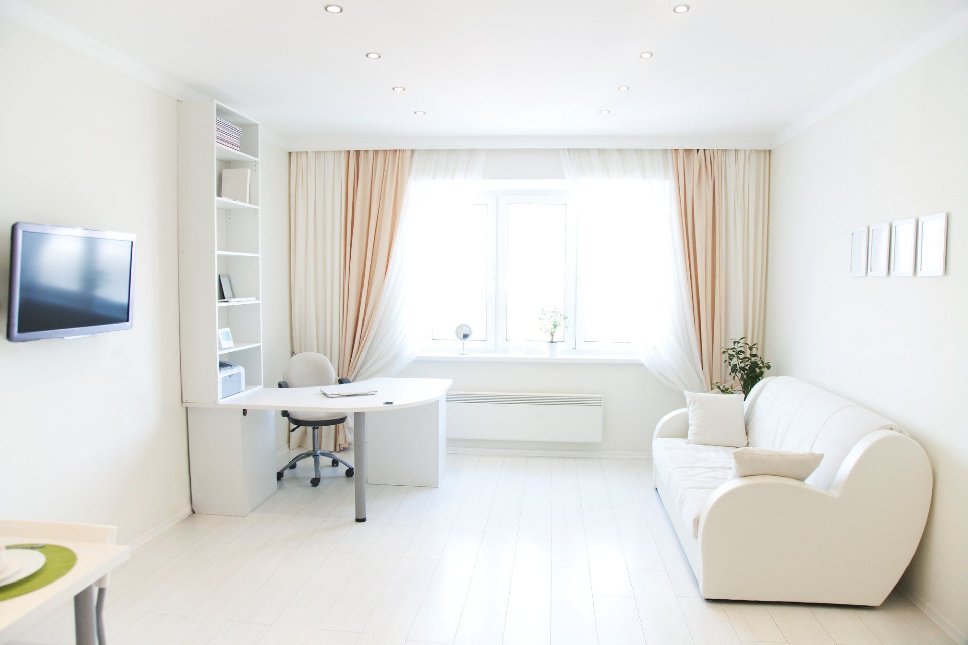 Modern room with white couch looking at wall mounted TV in foreground. Buld in desk with chair, framed by curtains pulled open on window with light flowing in.