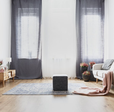 Long full length opaque grey curtains over loungeroom windows. Timber floored lounge with white couch, timber dresser and large grey rug.