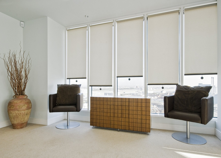 Modern lounge chairs either side of timber dresser in lounge with roller blinds covering 5 windows, at differnt lengths.