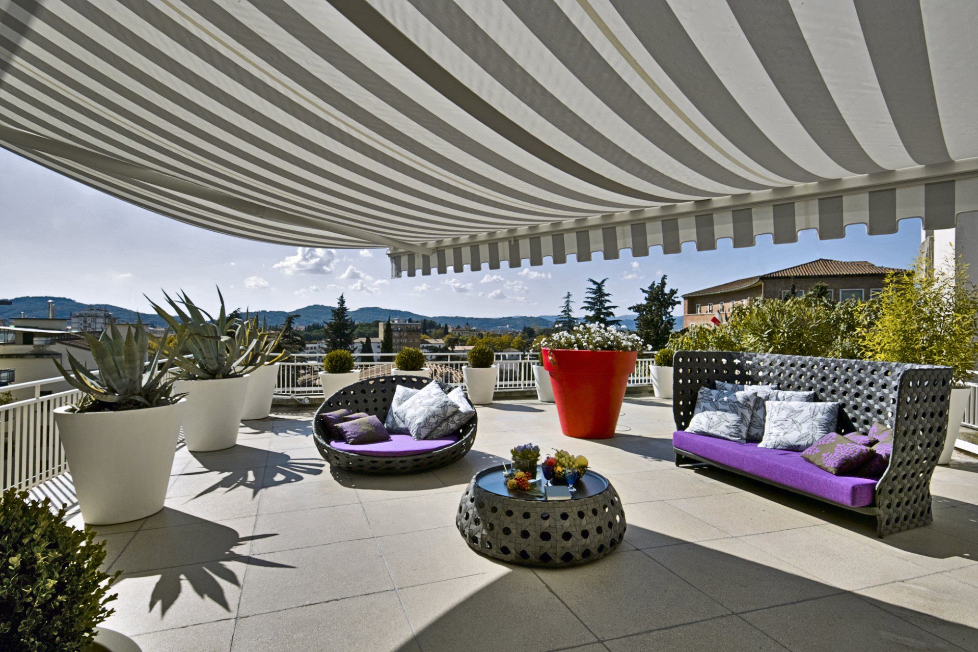 Outdoor awning extending out over outdoor living area with outdoor lounges and large potted plants in the background.