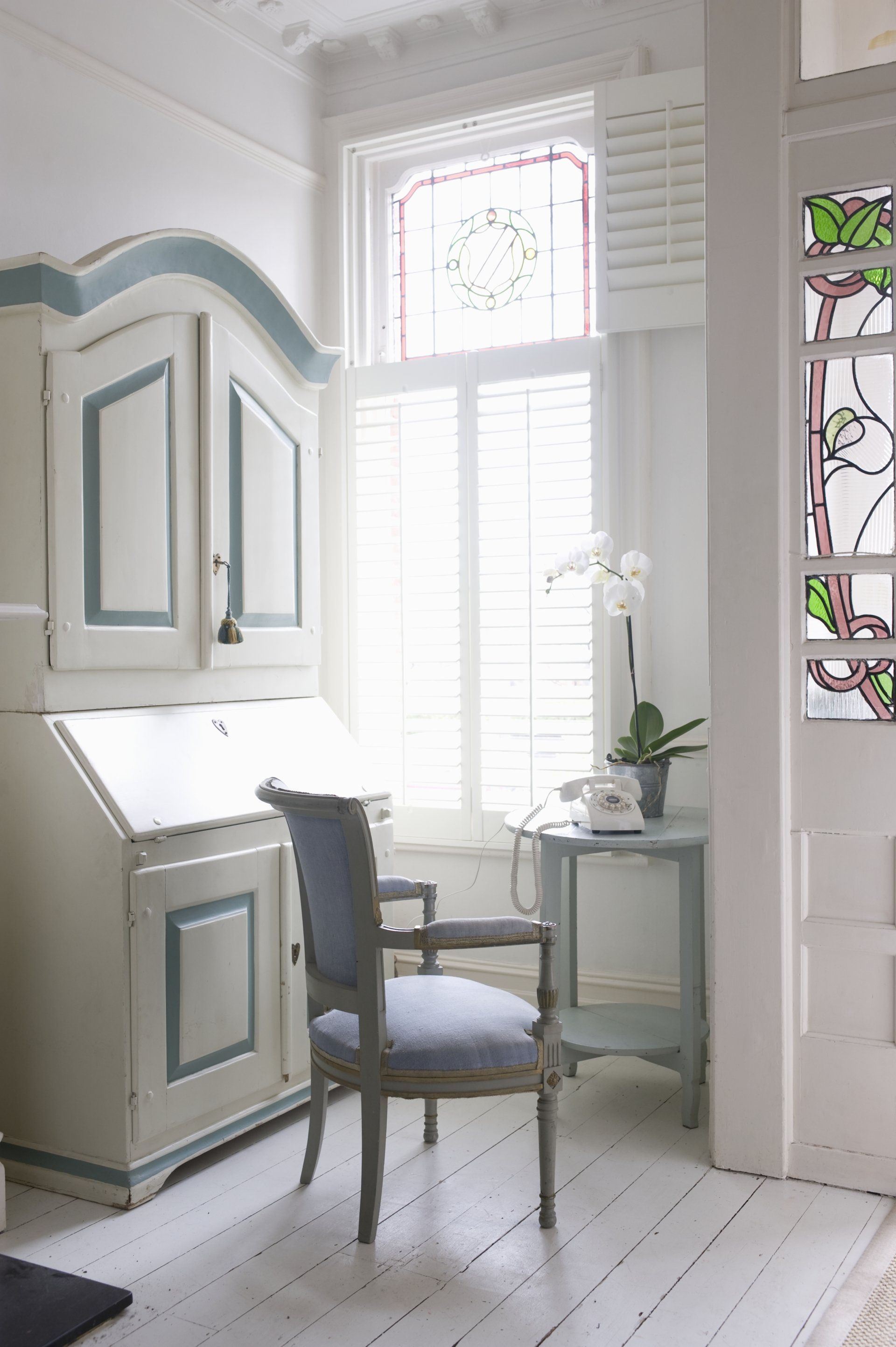 Light shining through open plantation shutters in period style house. In front of window is a chair, and table with a telephone and plant, adjacent to a large wooden cupboard.