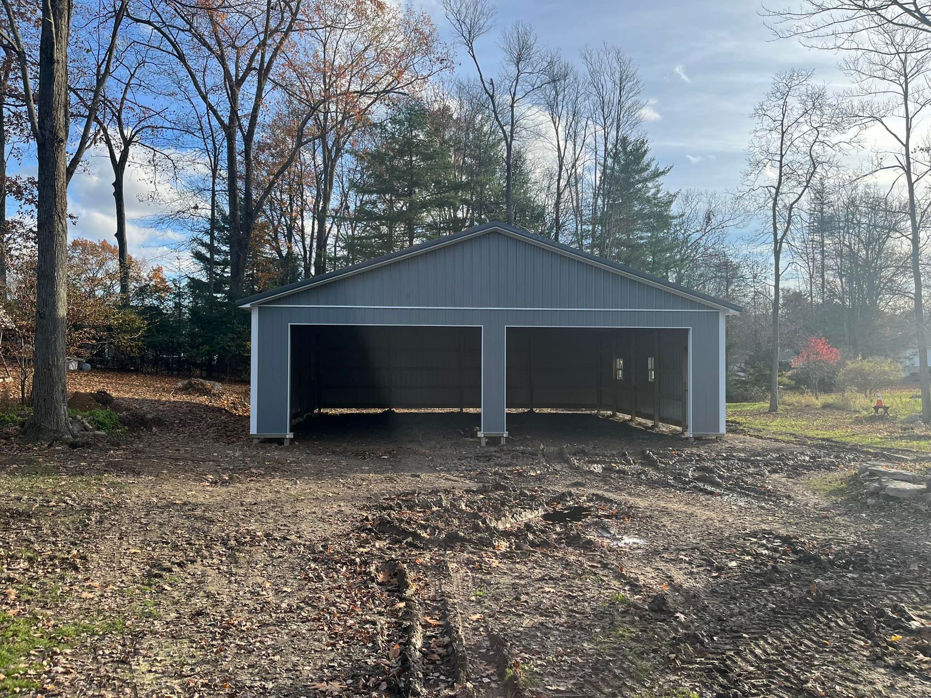 Gray detached two-bay garage in a wooded yard on a dirt driveway, with trees and autumn leaves around it