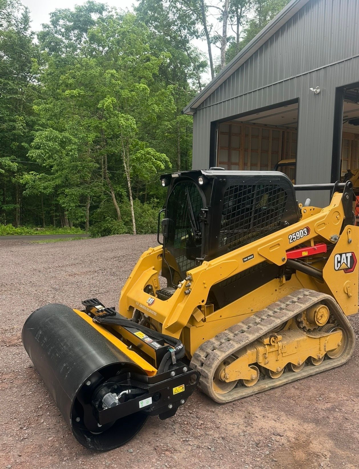 Yellow skid steer with a black roller attachment on gravel beside a gray building and trees.