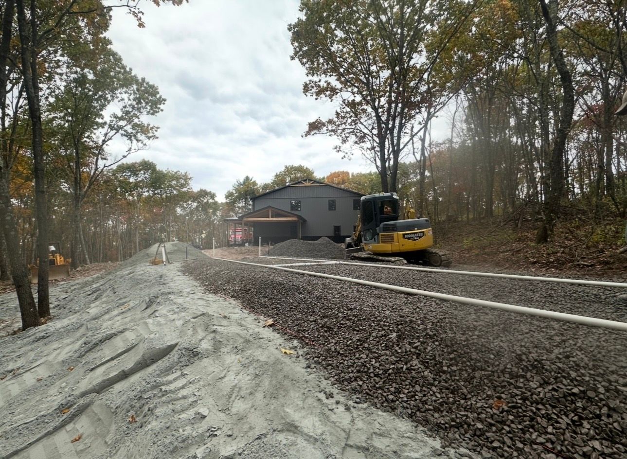 Construction site with yellow excavator beside a gray house and dirt road in a wooded area