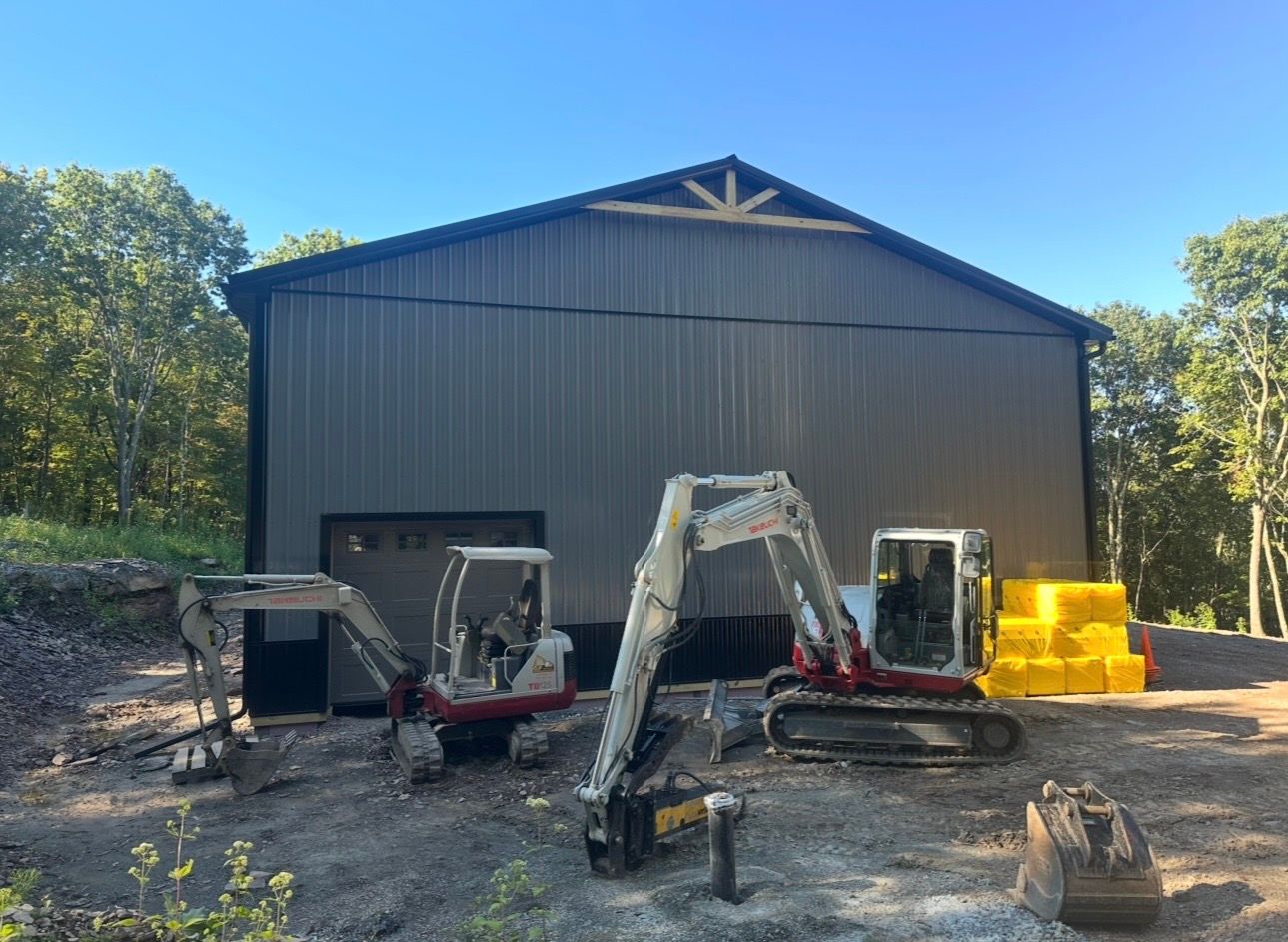 Large dark metal barn with three excavators parked outside on a gravel lot under a clear blue sky.