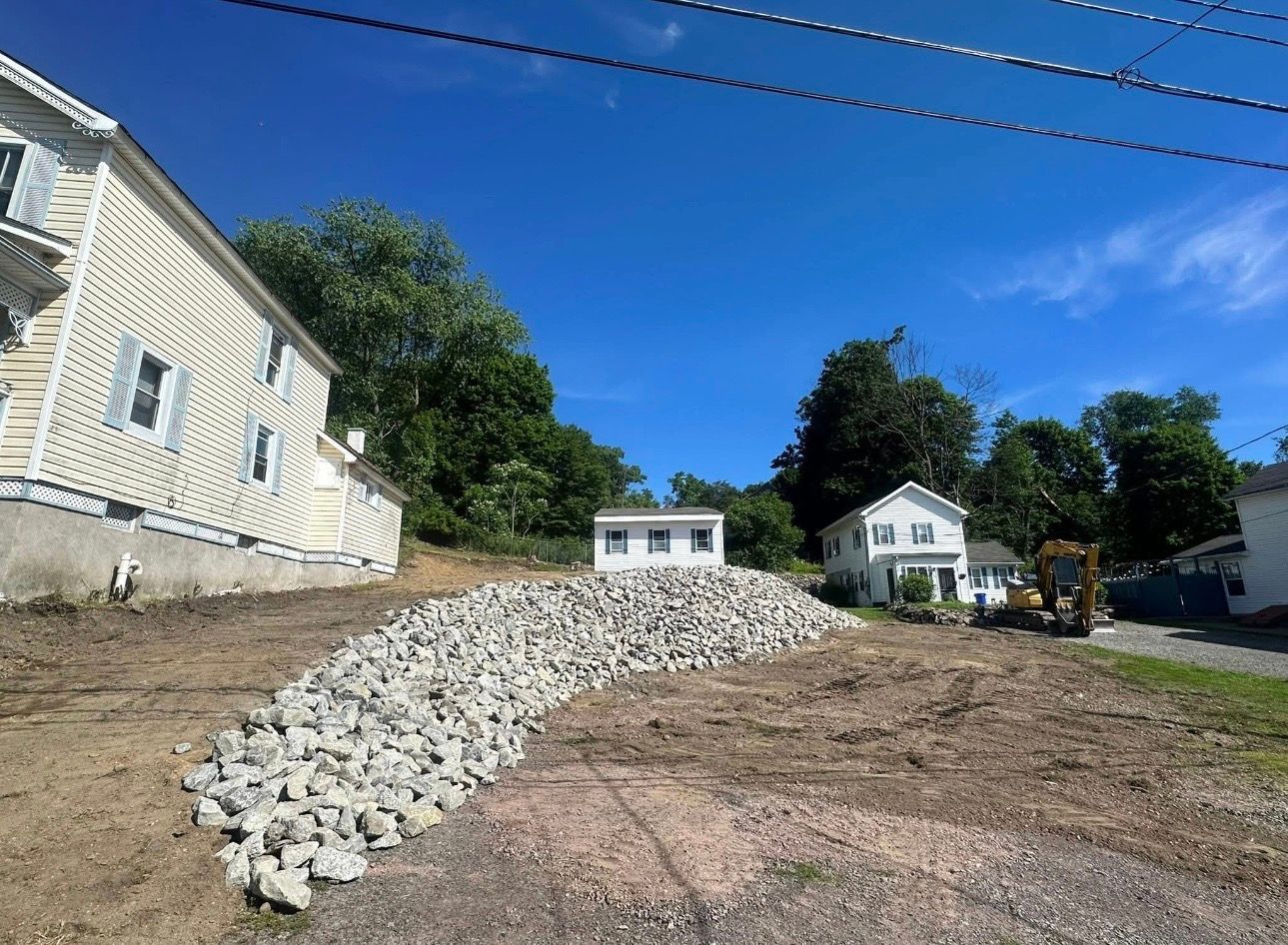 Rocky dirt lot with gravel mound, houses, trees, and a bright blue sky