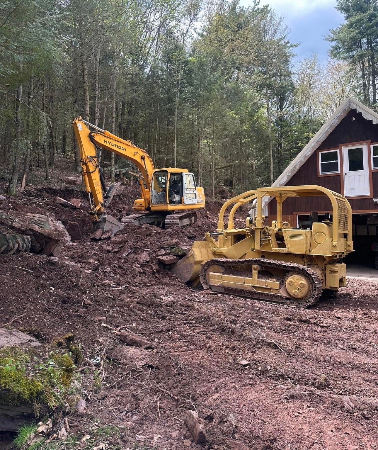 Yellow excavator and bulldozer on muddy ground beside a cabin in a wooded area