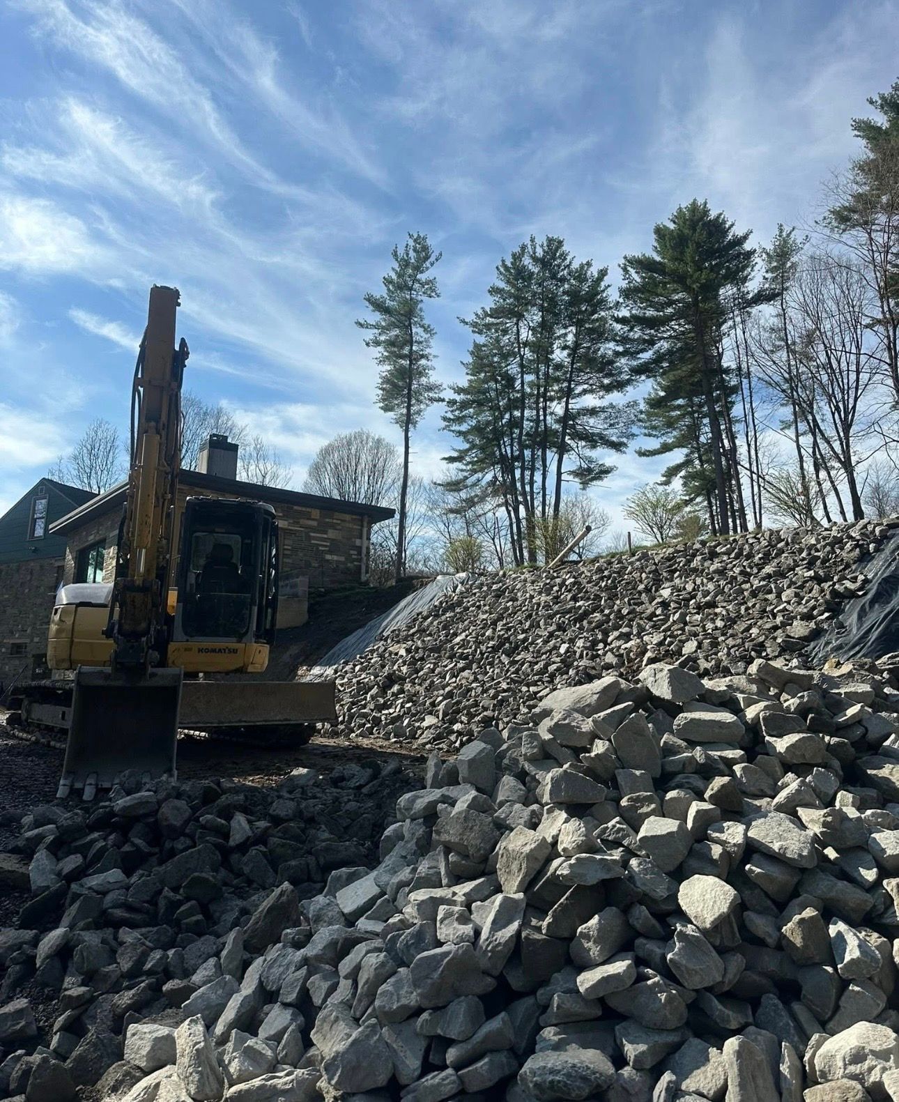 Excavator beside a large pile of gray rocks under a clear sky with trees in the background