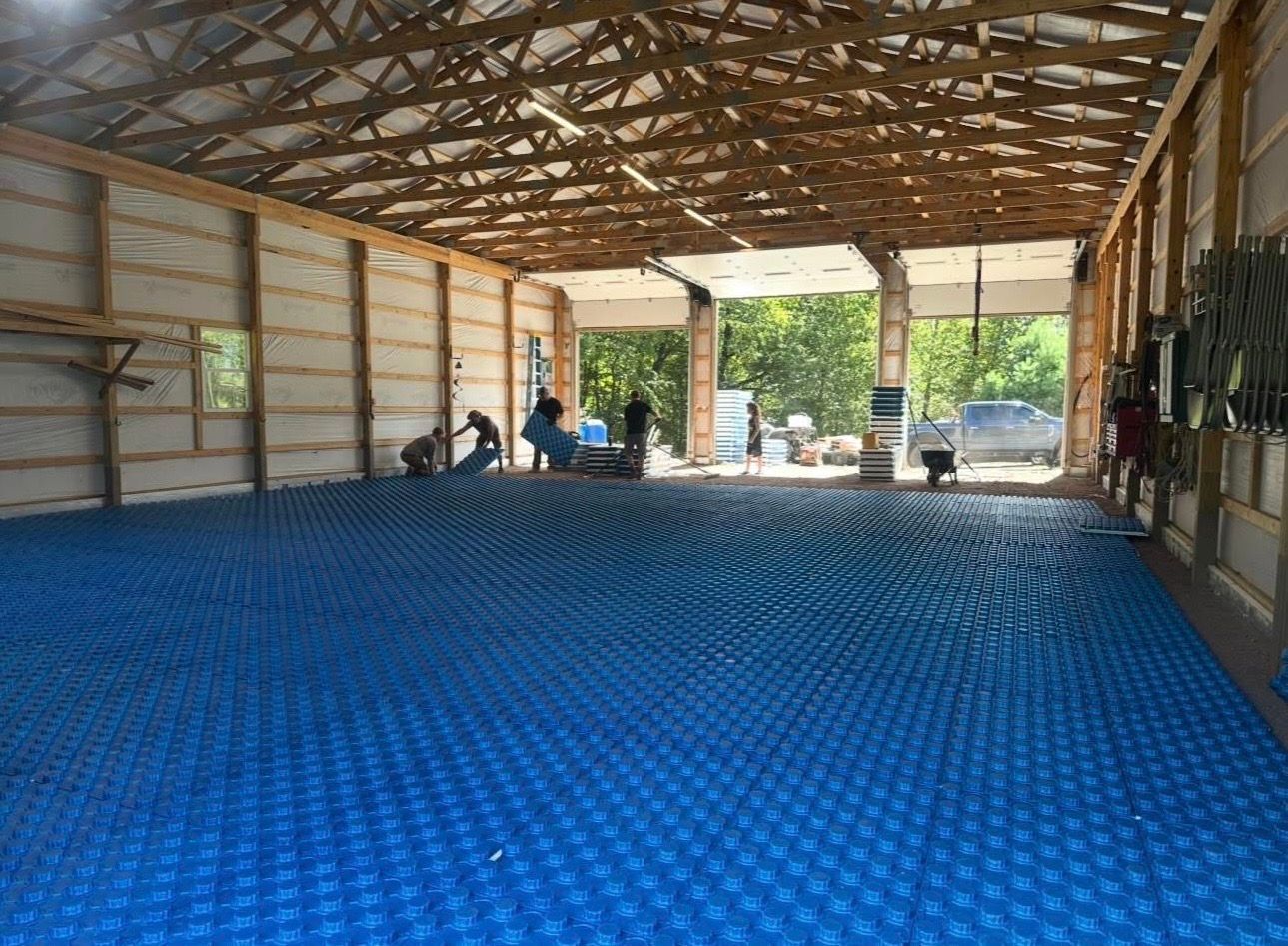 Large barn interior with blue patterned floor, wooden roof beams, and a few people near the open entrance.