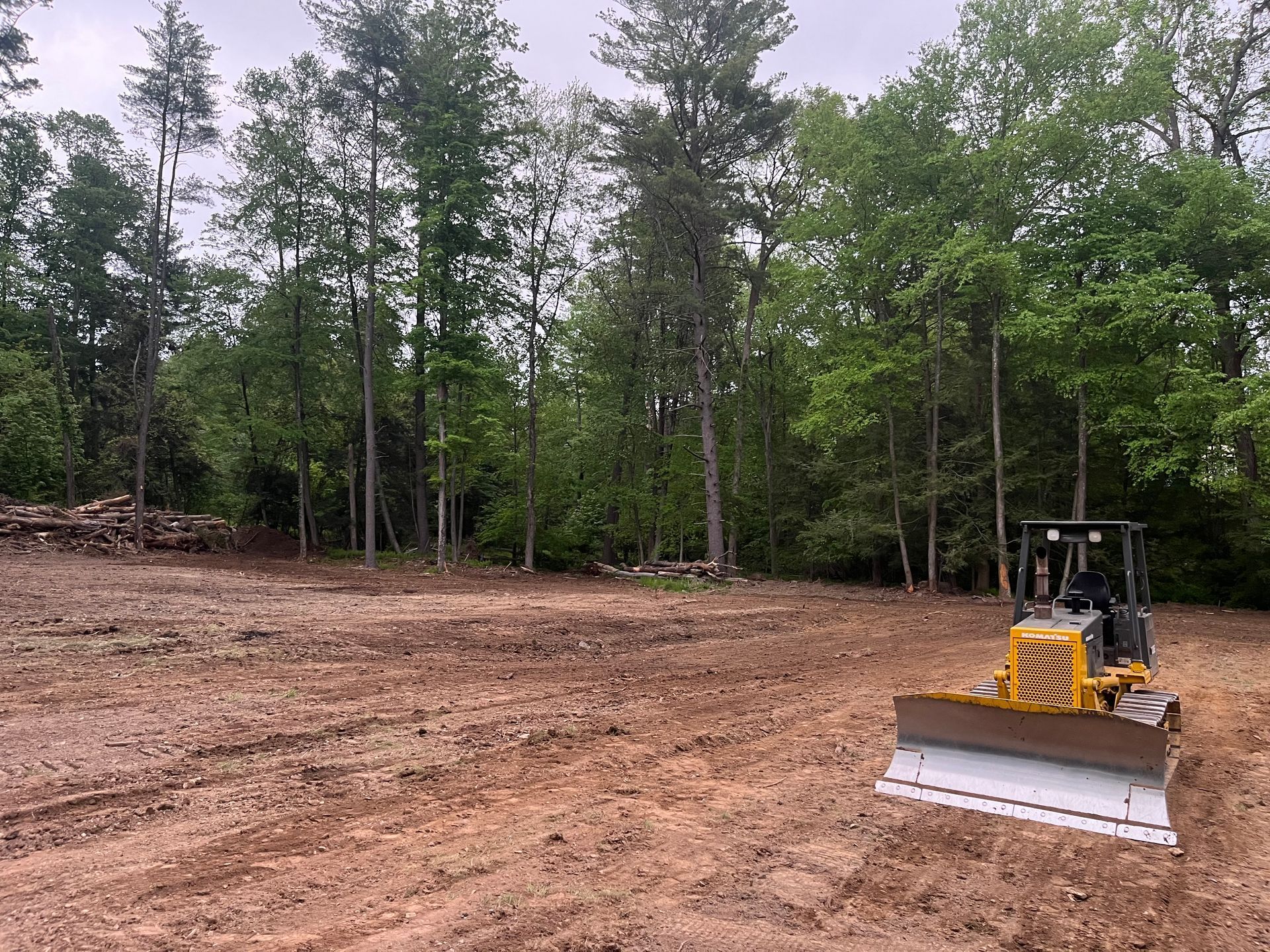 Bulldozer grading a dirt lot beside a tree line under a cloudy sky