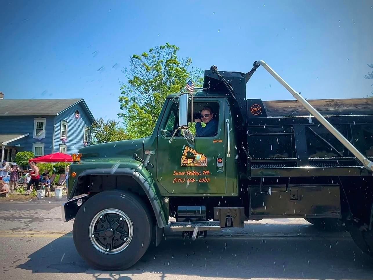 Green dump truck parked at a parade, with driver visible in the cab and spectators in the background.