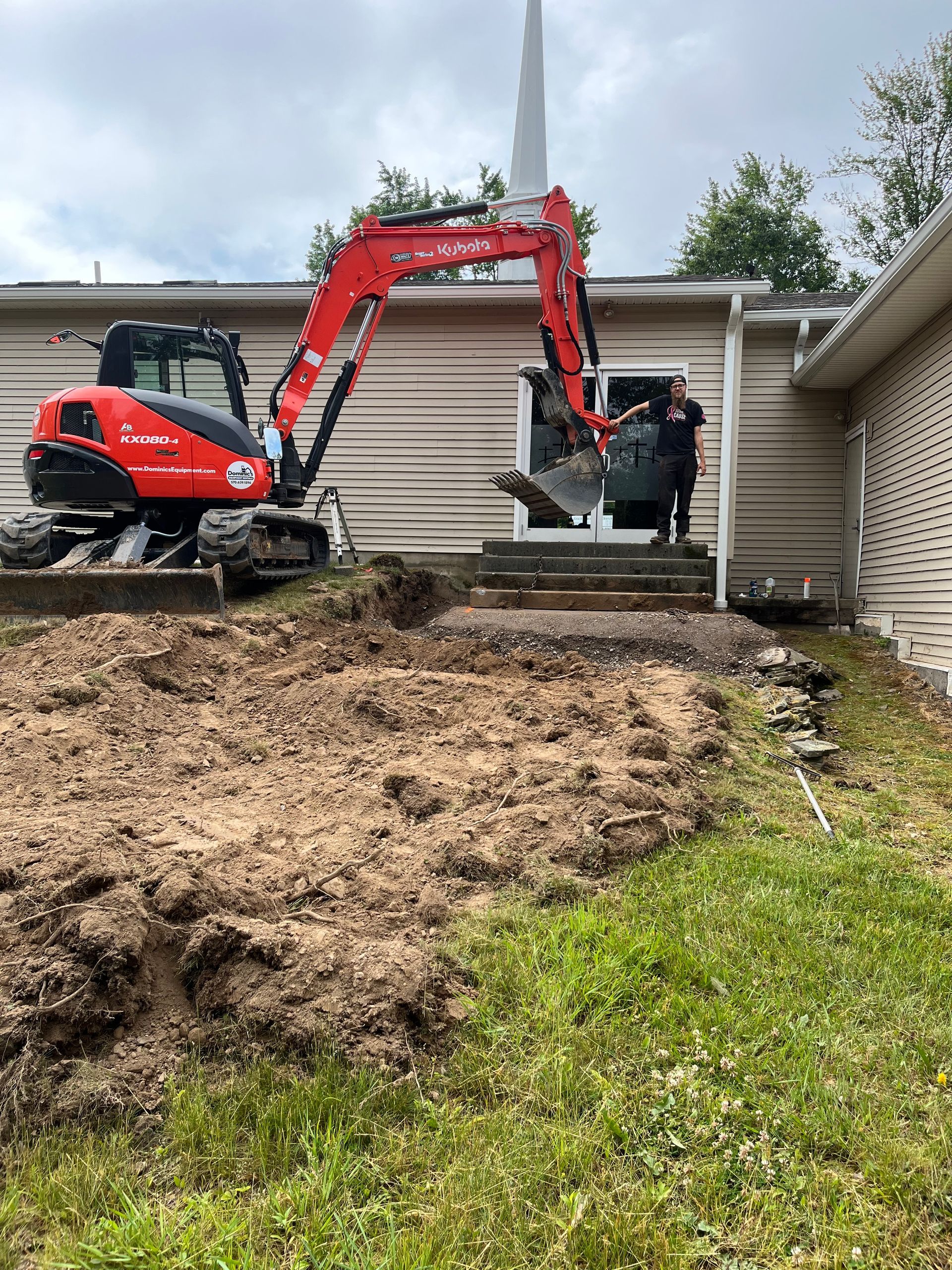Red excavator lifting a worker beside a house during yard excavation.
