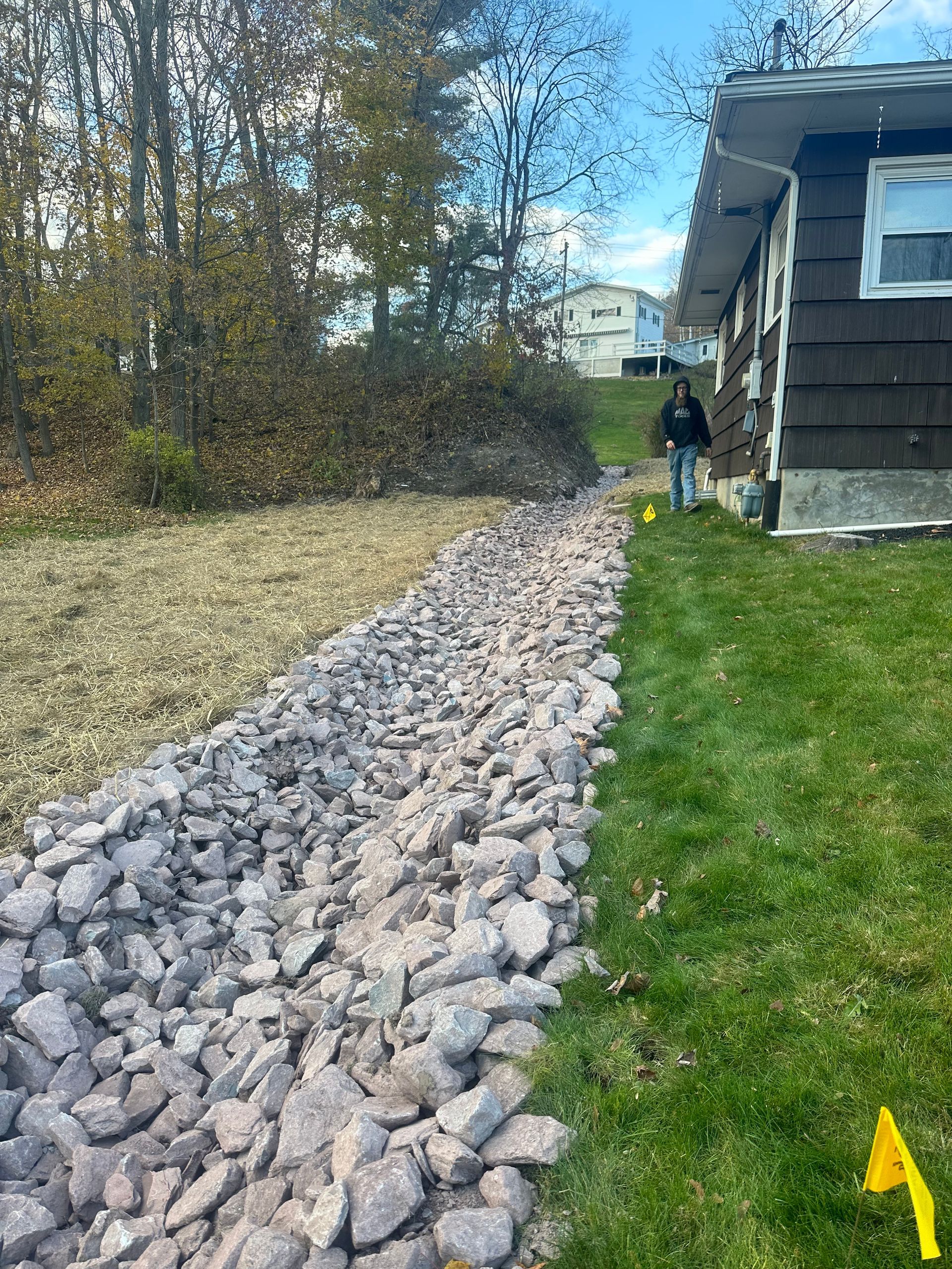 Rock-lined path beside a house, with a person standing in the distance and a yellow flag marker on the grass.