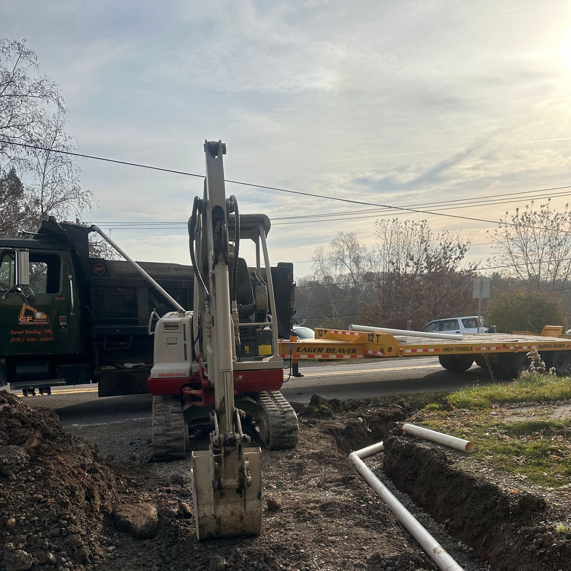 Excavator at a muddy worksite beside a truck, with pipes and dirt piles under a cloudy sky