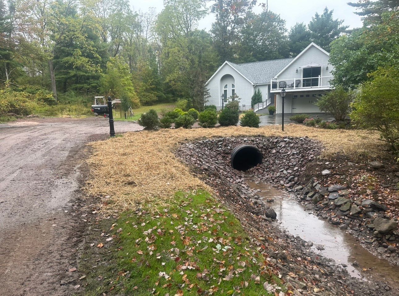 White house by a gravel drive and culvert beside a wet ditch in a wooded yard.