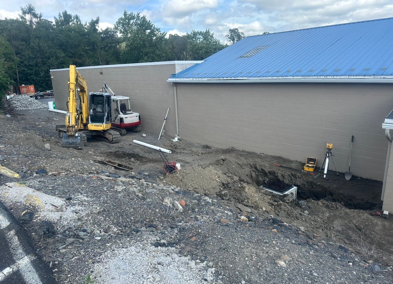 Construction site beside a gray building with a blue roof and an excavator digging along the foundation