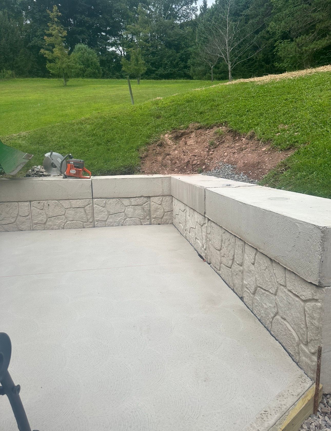 Concrete patio with low stone wall beside a grassy yard and dirt slope; red tool on the left.