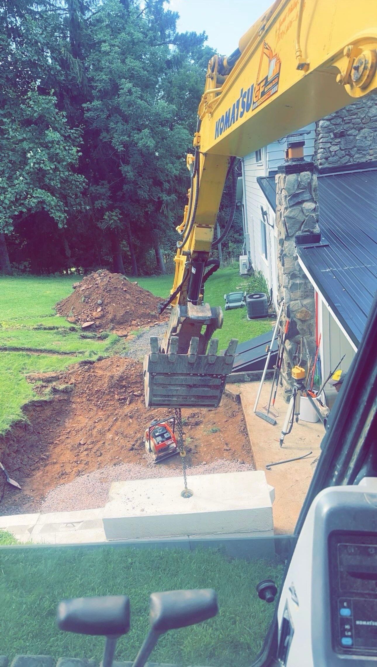 Yellow excavator digging soil beside a house, with a dirt pile and green lawn nearby.