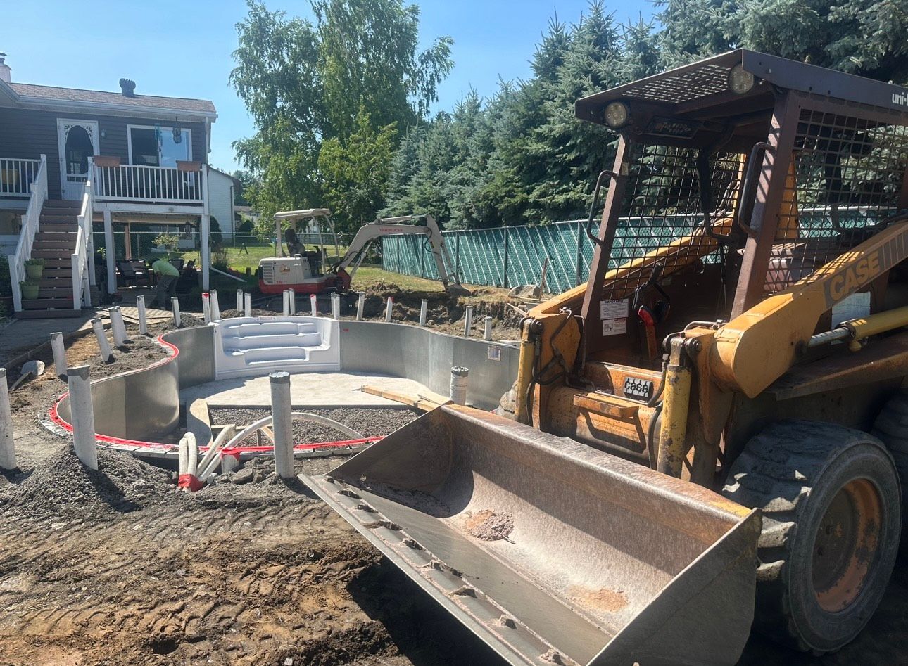 Backyard construction scene with a skid steer, concrete forms, and a house in the background