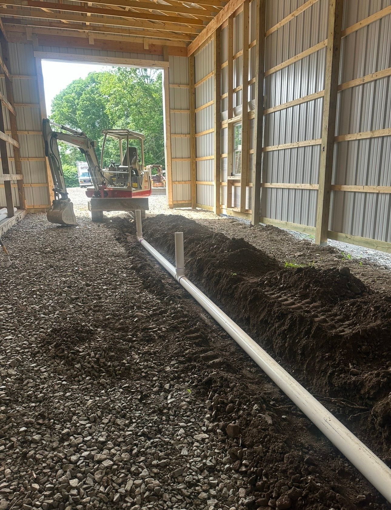 Barn interior with gravel floor, white pipe, and excavator near an open end showing trees outside