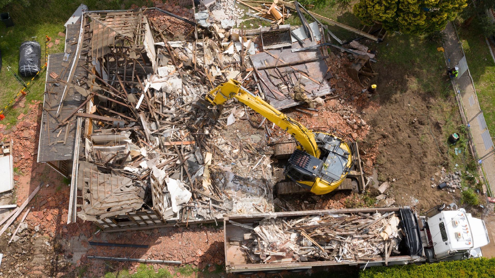 An aerial view of a house being demolished with a yellow excavator.