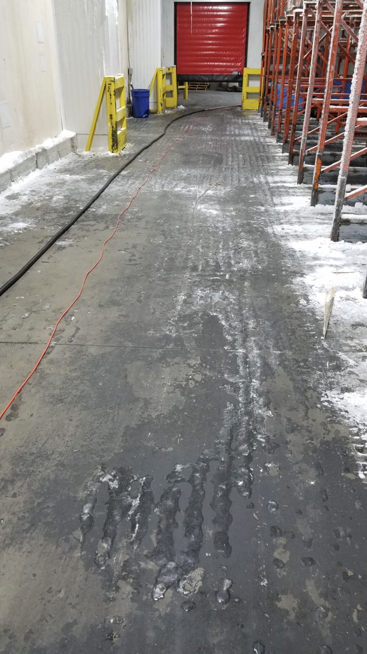 Warehouse floor with patches of water and possible ice, leading to a closed red door. Yellow safety blocks are in view.