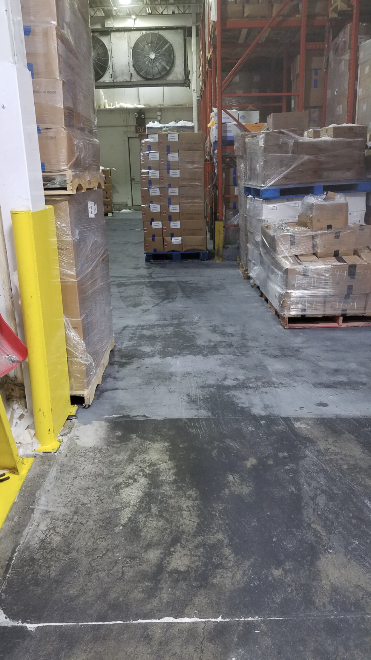 Warehouse aisle with stacked boxes on pallets, cold storage unit visible in background, wet floor.