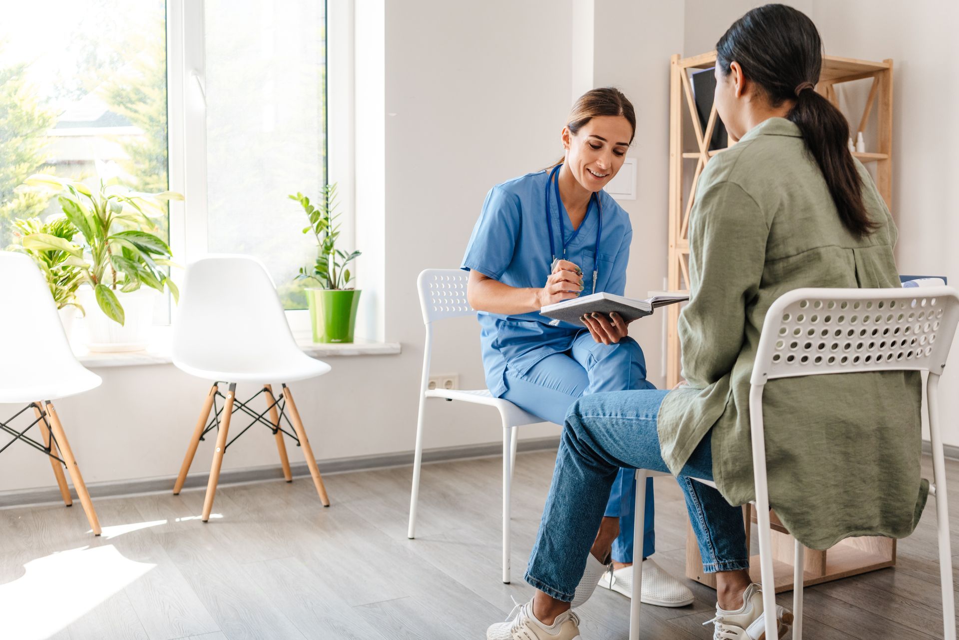 Two women talking at a table in a bright office, one taking notes while the other listens.