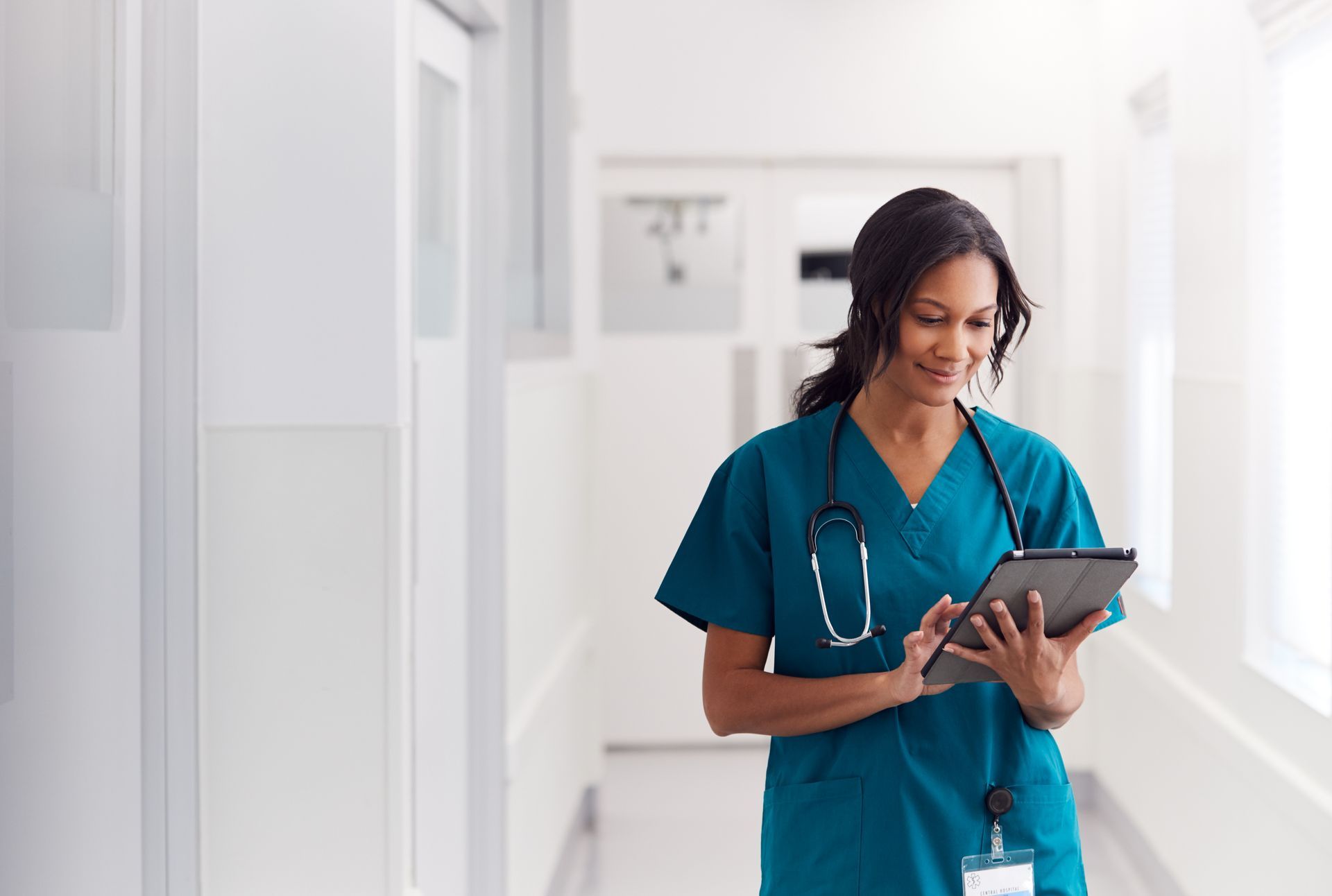 Nurse in teal scrubs smiling while using a tablet in a bright hospital hallway