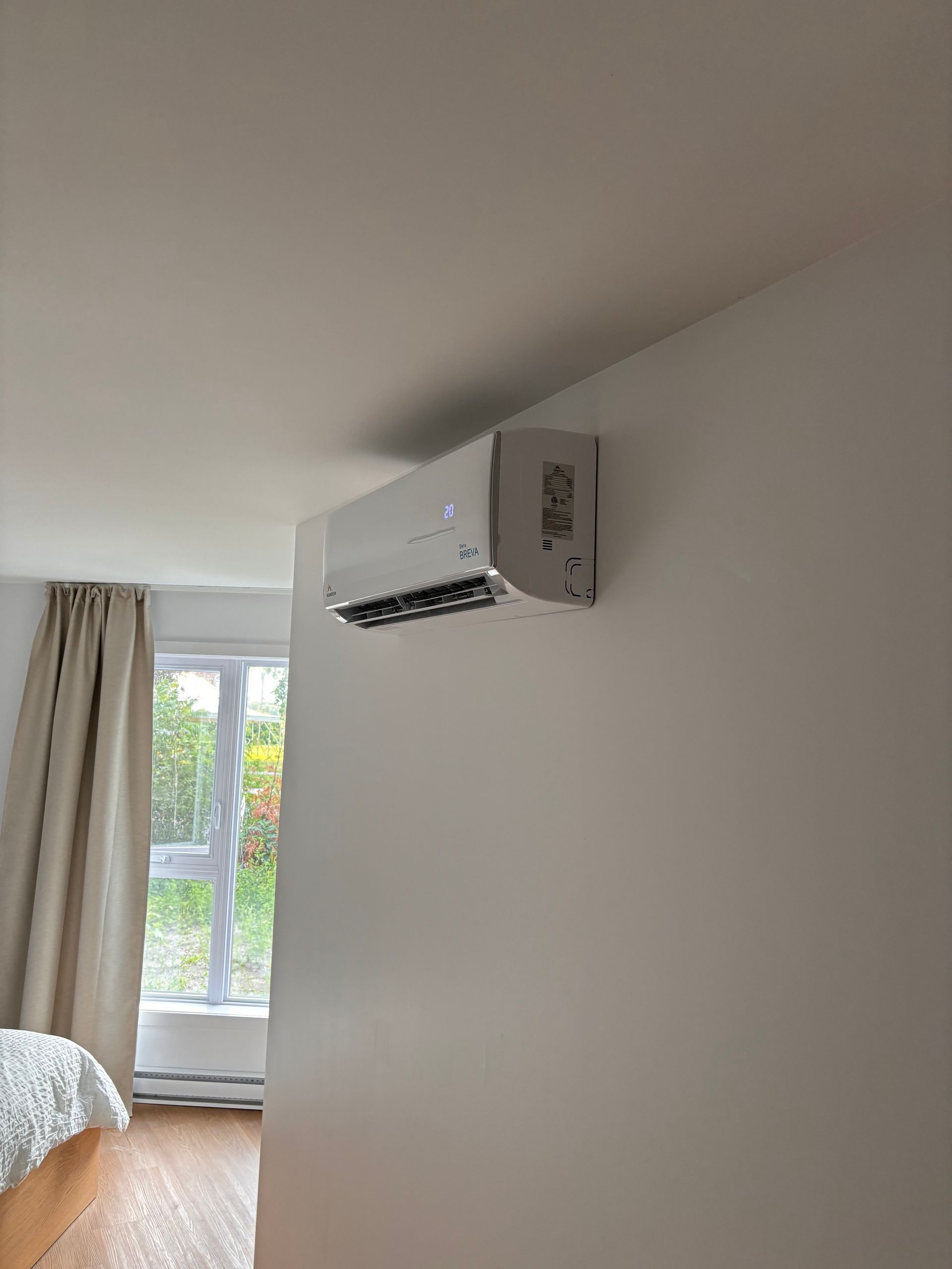 White wall-mounted air conditioner installed in a bedroom with window and beige curtain.