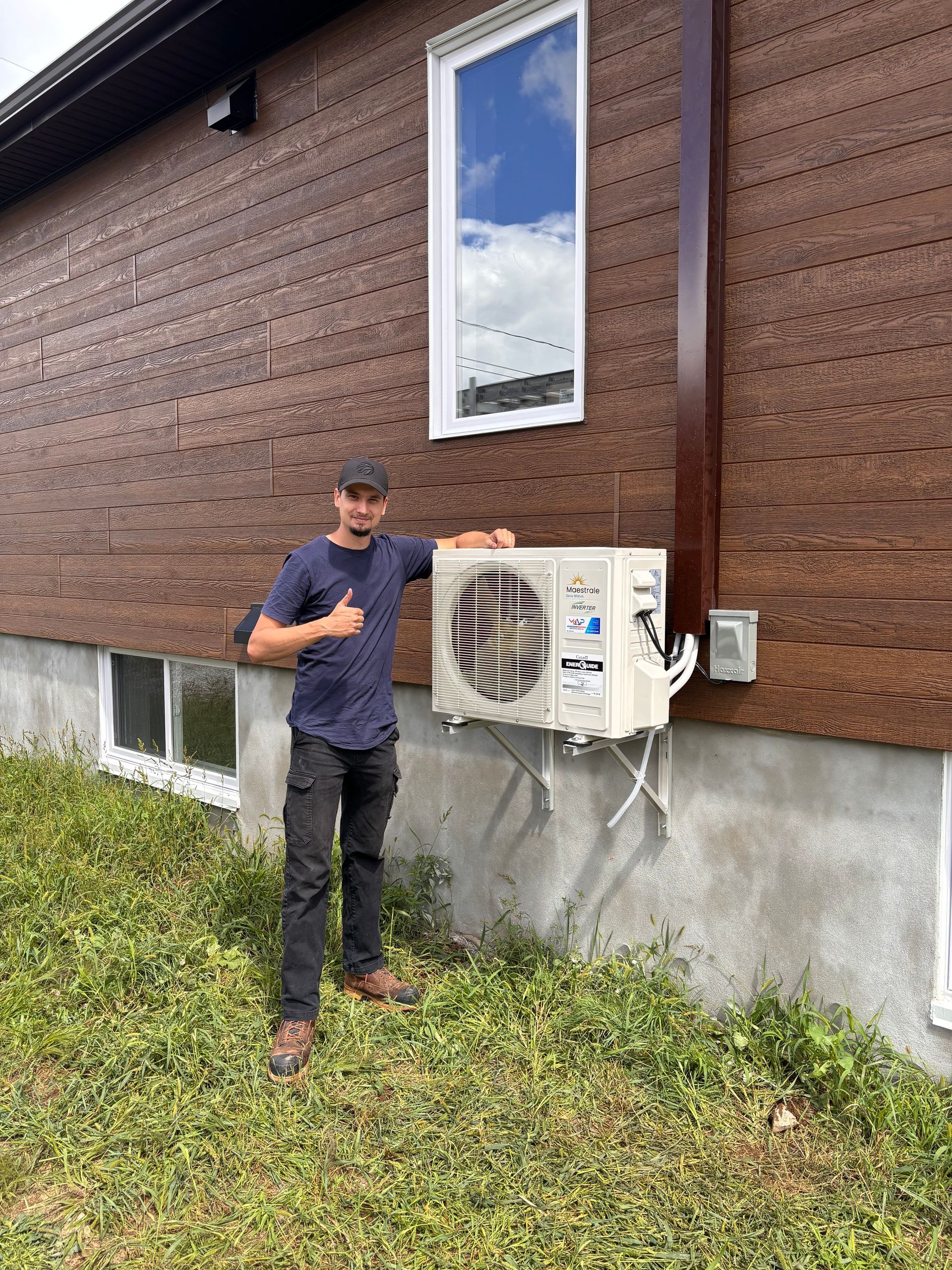 Un homme se tient à côté d'un climatiseur sur un bâtiment aux murs bruns et lève le pouce.